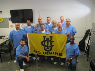 Group of inaugural UCI LIFTED students stand behind gold and blue UC Irvine banner in classroom at Richard J. Donovan Correctional Facility.